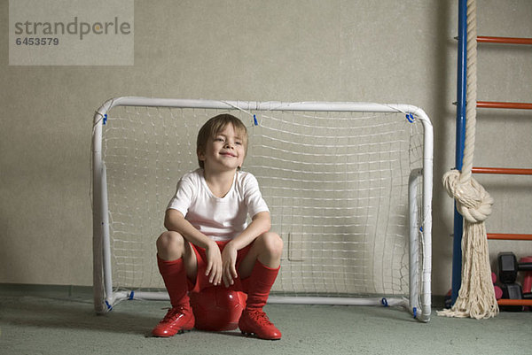 Ein Junge sitzt auf einem Fußball vor einem Fußballtor.