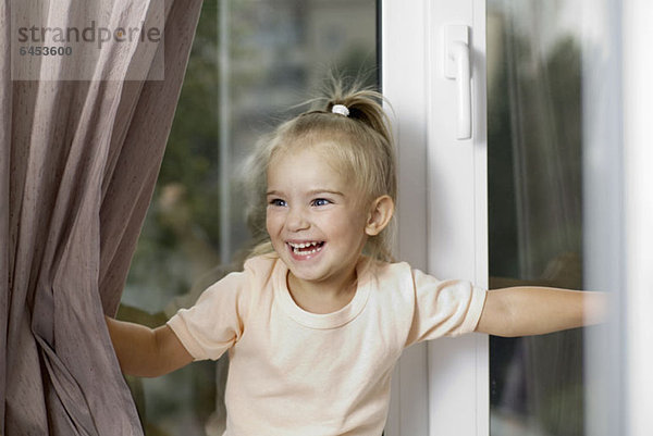 Ein junges lachendes Mädchen schiebt Fenstervorhänge auseinander.