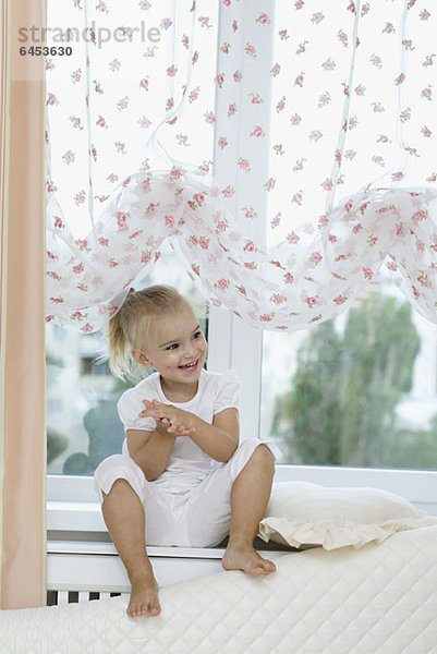 Ein junges Mädchen sitzt auf einem Fensterbrett und klatscht aufgeregt in die Hände.