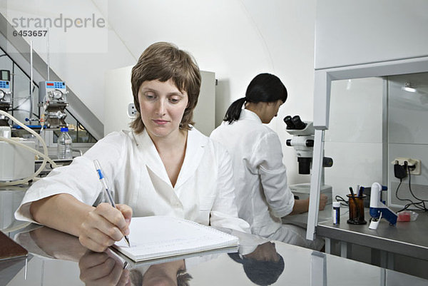 Ein Laborant schreibt Notizen  während ein anderer Techniker im Hintergrund arbeitet.