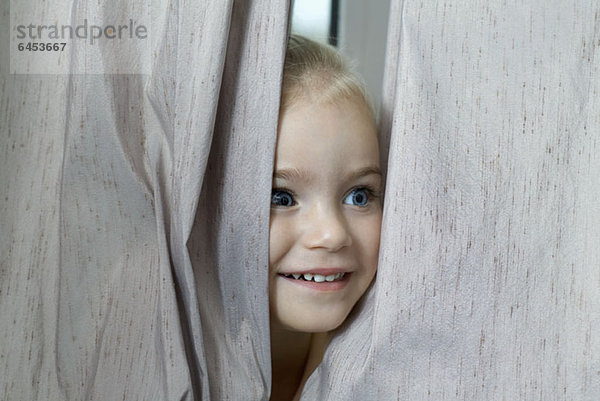 Ein junges Mädchen  das sich zwischen Fenstervorhängen versteckt.