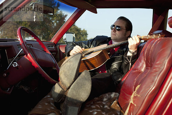 Ein Rockabilly  der in seinem Oldtimer eine Akustikgitarre spielt.