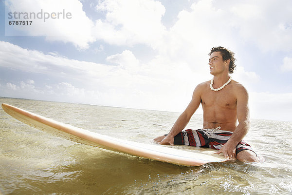Hawaii  Oahu  junger Mann sitzt auf seinem Surfbrett an der Küstenlinie.