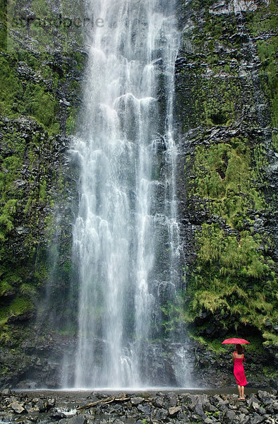 Hawaii  Maui  Kipahulu  Hana  Frau steht mit Sonnenschirm am Fuße des Wasserfalls.