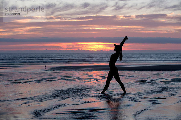 Silhouette einer jungen Frau  die sich bei Sonnenuntergang am Meer ausstreckt.