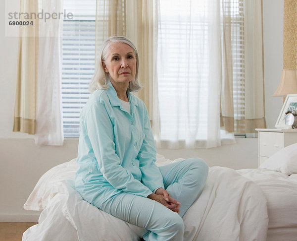 Porträt einer traurigen älteren Frau  die im Schlafanzug auf dem Bett sitzt.