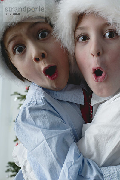 Jungen mit Weihnachtsmützen  die sich umarmen und überraschte Gesichter machen.