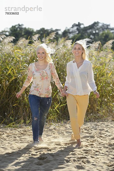 Zwei fröhliche blonde junge Frauen gehen Hand in Hand an einem Sandstrand