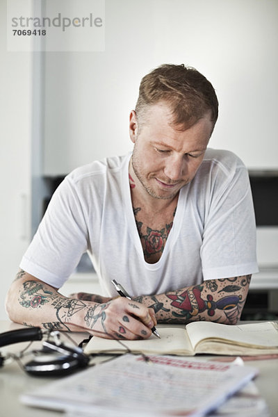 Tätowierter Mann beim Schreiben im Buch am Schreibtisch