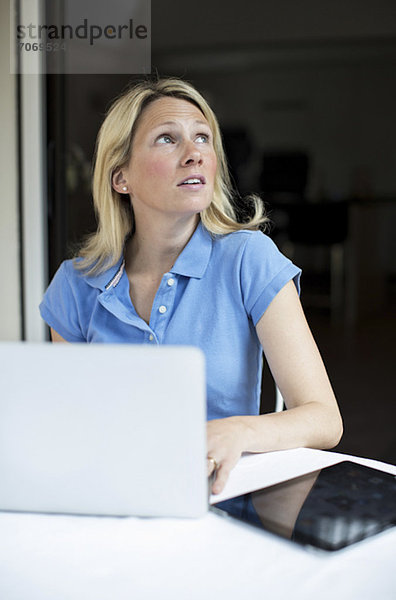 Mittlere erwachsene Frau sitzend mit Laptop und digitalem Tablett  während sie nach oben schaut.