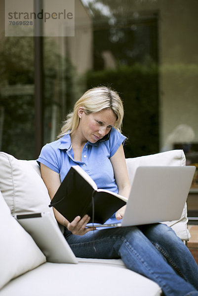 Mittlere erwachsene Frau mit Laptop und Handy  die ein Buch im Wohnzimmer hält.