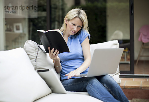 Mittlere erwachsene Frau mit Laptop und Handy  die ein Buch im Wohnzimmer hält.