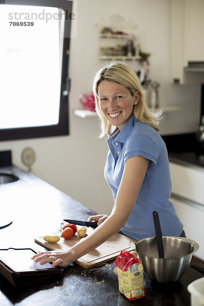 Mittlere erwachsene Frau mit Rezept auf digitalem Tisch und Zubereitung des Essens in der Küche
