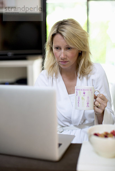 Mittlere erwachsene Frau im Bademantel mit Laptop beim Kaffeetrinken