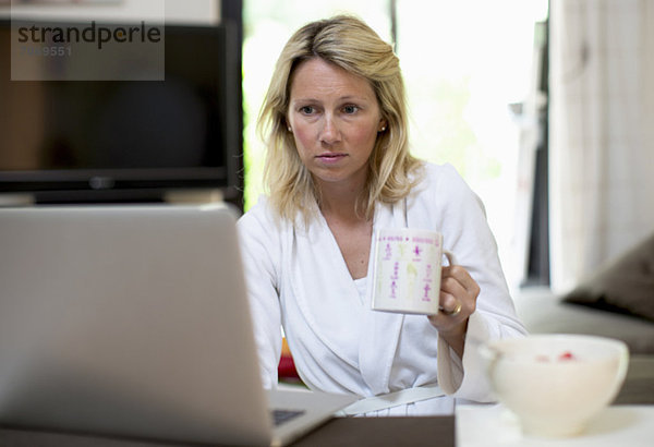 Mittlere erwachsene Frau im Bademantel mit Laptop beim Kaffeetrinken