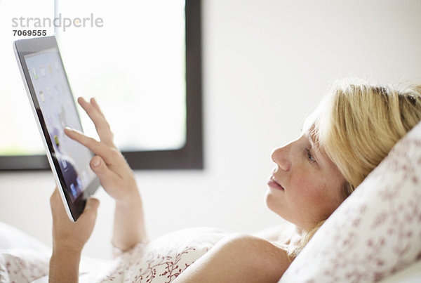 Lässige  erwachsene Frau  die auf dem Bett liegend auf ein digitales Tablett schaut.