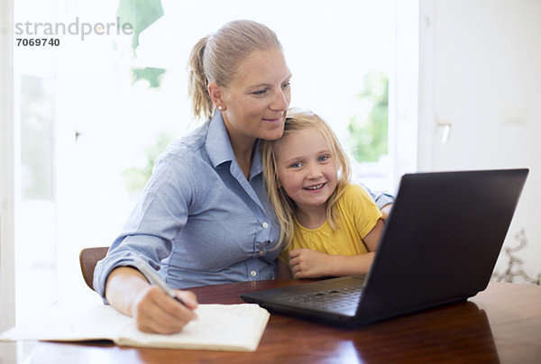 Mid Erwachsene Frau mit Laptop beim Schreiben von Notizen sitzen mit Tochter am Schreibtisch