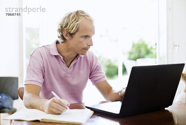 Mid Erwachsene Mann mit Laptop beim Schreiben von Notizen am Schreibtisch