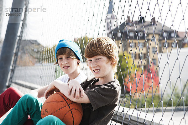 Zwei Jungen sitzen mit Basketball an einem Zaun