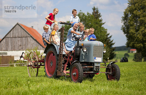 Frau mit Kindergruppe auf altem Traktor vor dem Bauernhaus sitzend