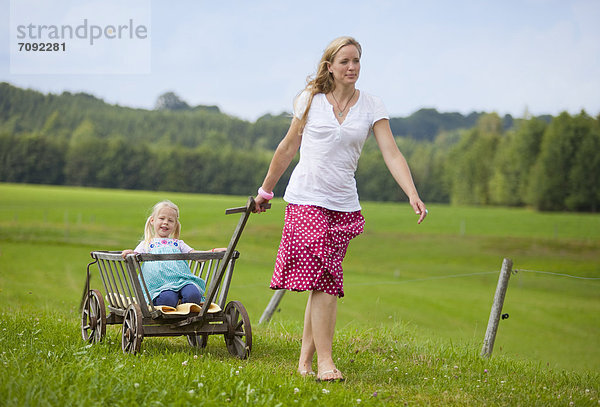 Germany  Bavaria  Mother pulling daughter in hand cart through meadow