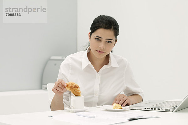 Junge Frau taucht Croissant in Kaffee am Schreibtisch im Büro ein