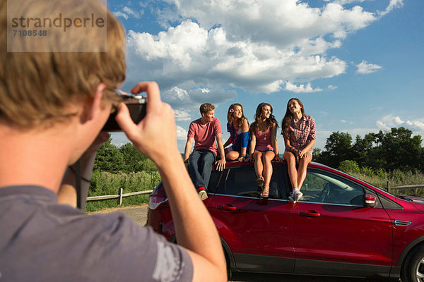 Freunde sitzen auf dem Autodach  junger Mann fotografiert