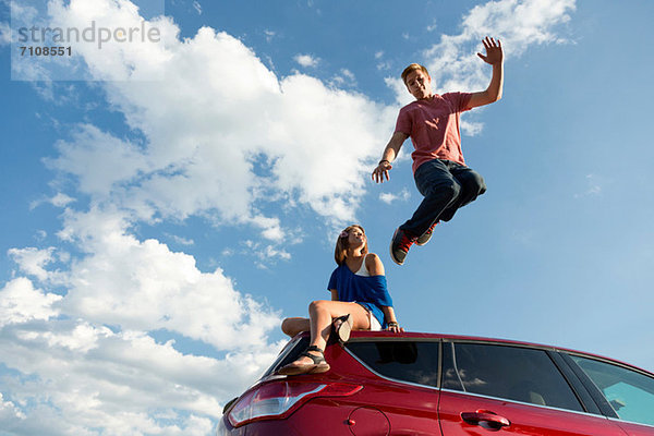Junger Mann springt vom Autodach  Mädchen sitzt auf dem Dach