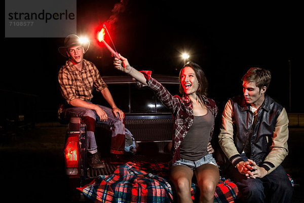 Drei Freunde sitzen nachts auf der Heckklappe des Autos  ein Mädchen hält eine Wunderkerze.