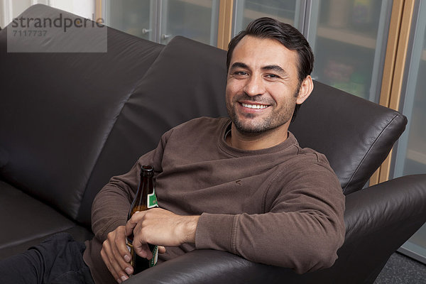 Mann sitzt lächelnd mit Bierflasche auf einem Ledersofa