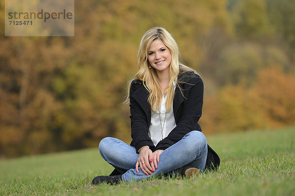 Lächelnde blonde junge Frau sitzt auf herbstlicher Wiese