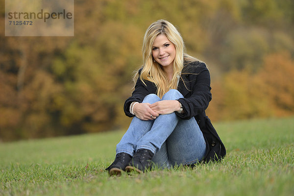 Lächelnde blonde junge Frau sitzt auf herbstlicher Wiese