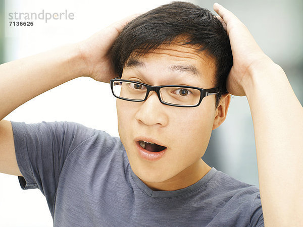 Junger Mann  Asiate  Student mit Brille  erstaunt  Hände hinter Kopf verschränkt