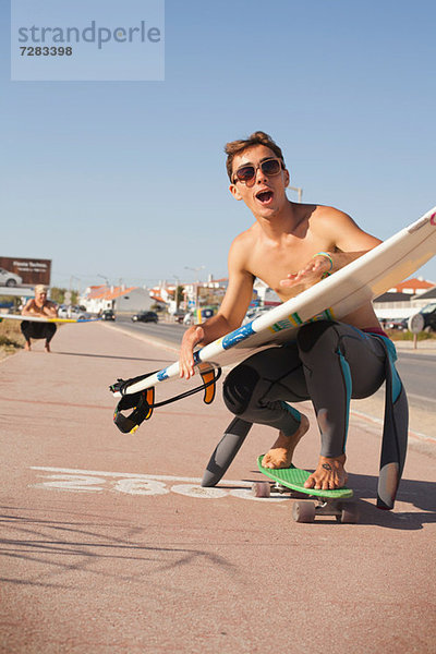 Junger Mann beim Skateboarden auf der Straße  während er ein Surfbrett hält.