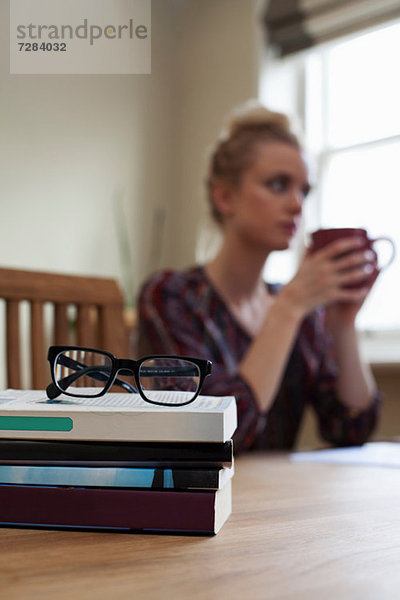 Bücher und Brille im Vordergrund  junge Frau mit Kaffeetasse im Hintergrund