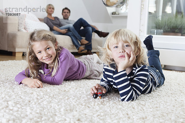 Junge und Mädchen liegen auf dem Boden  die Eltern sitzen auf der Couch im Hintergrund.