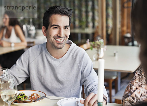 Glücklicher Mann  der eine Freundin am Restauranttisch mit Menschen im Hintergrund ansieht.