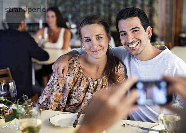 Glückliche junge Freunde  die am Restauranttisch mit Menschen im Hintergrund fotografieren.
