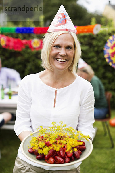 Porträt einer glücklichen erwachsenen Frau mit einem Teller voller gekochtem Hummer und Freunden im Hintergrund.