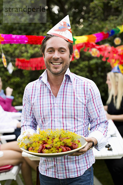 Porträt eines glücklichen erwachsenen Mannes mit einem Teller voller gekochtem Hummer und Freunden im Hintergrund.