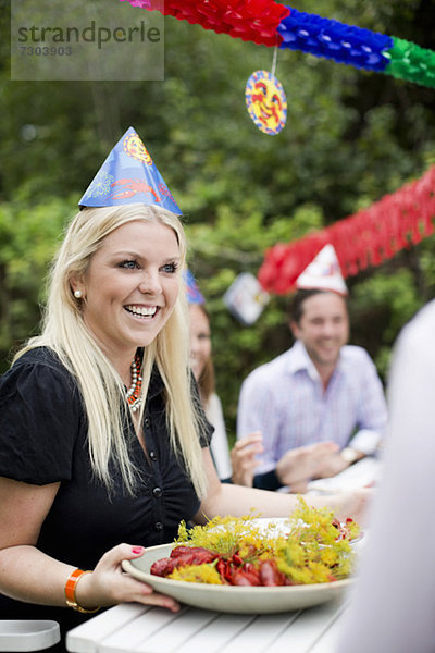 Fröhliche junge Frau  die am Esstisch während der Krebsparty an einem Teller voller gekochter Hummer vorbeigeht.