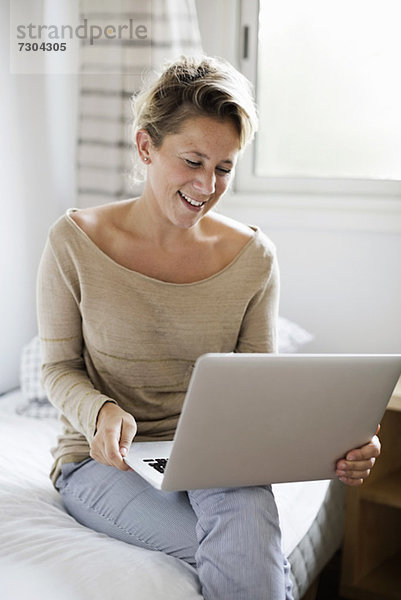 Glückliche mittlere erwachsene Frau mit Laptop im Bett zu Hause
