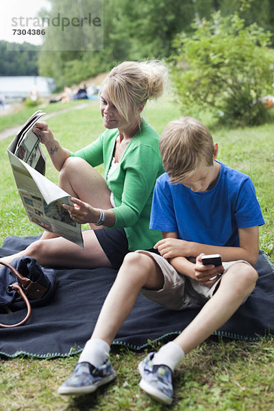 Reife Frau liest Zeitung sitzend mit Sohn mit Handy im Park