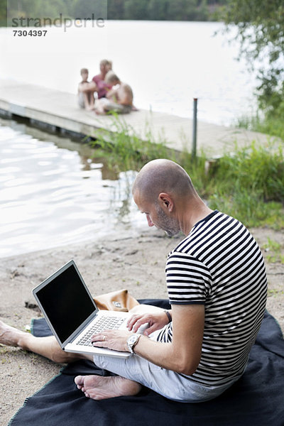 Erwachsener Mann mit Laptop am Strand mit Familie auf dem Pier sitzend