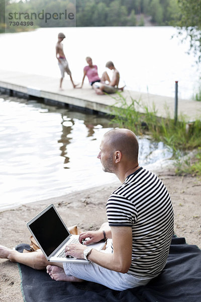 Der reife Mann schaut weg  während er den Laptop am Strand benutzt und die Familie am Pier sitzt.