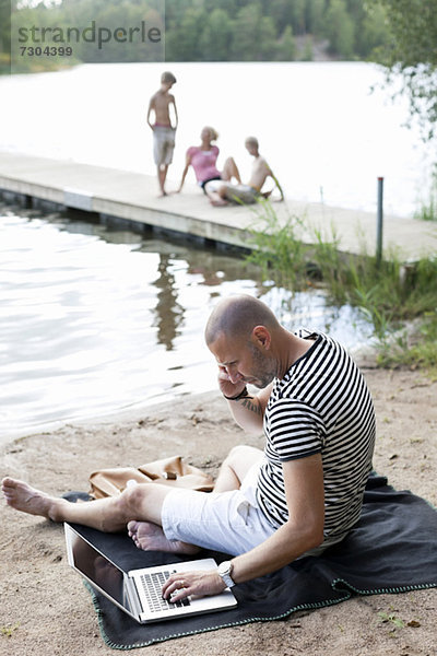 Reifer Mann mit Laptop bei der Kommunikation auf dem Handy am Strand mit der Familie am Pier sitzend