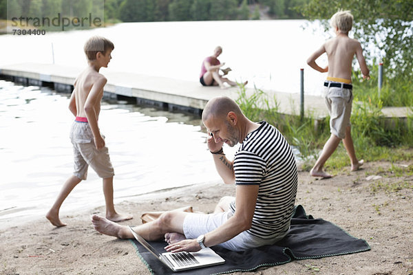 Reifer Mann mit Laptop bei der Kommunikation auf dem Handy am Strand mit der Familie im Hintergrund