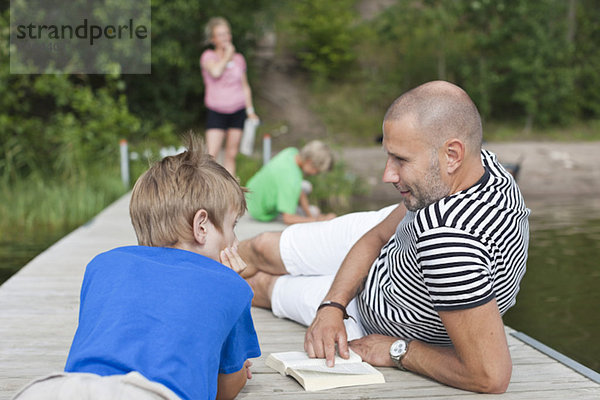 Erwachsener Mann mit Sohn auf dem Pier liegend und Familie im Hintergrund stehend