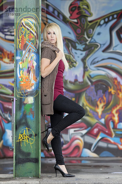 Junge Frau mit langen blonden Haaren und Jacke mit Kapuze posiert an mit Graffiti bemalter Stahlträger und vor Wand mit Graffiti