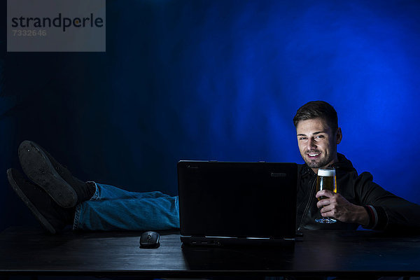 Junger Mann sitzt im Dunkeln vor seinem Laptop  trinkt ein Bier  Symbolbild Computersucht und Alkoholsucht